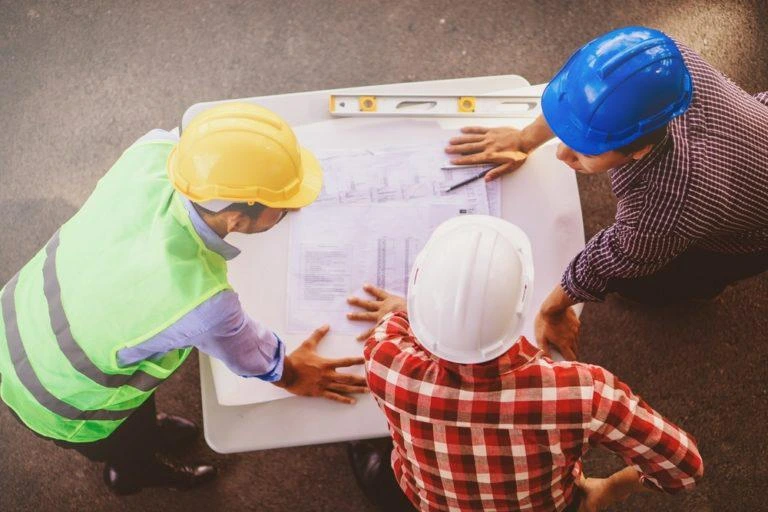 "Three men in construction helmets reviewing blueprints at a job site, representing field installers, site foreman, and project manager in the millwork industry."